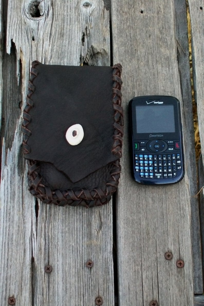 Op de afbeelding: Een bruine leren tas met een gevlochten rand en een bottenknop. De tas ligt naast een zwarte mobiele telefoon met een QWERTY-toetsenbord en de tekst "Verizon" en "Pantech" erop.