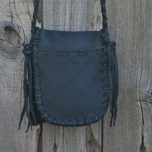 K&ouml;nnte beinhalten: Eine schwarze Ledertasche mit geflochtenem Trageriemen und Fransen. Die Tasche hat einen Klappverschluss und h&auml;ngt an einer Holzoberfl&auml;che.