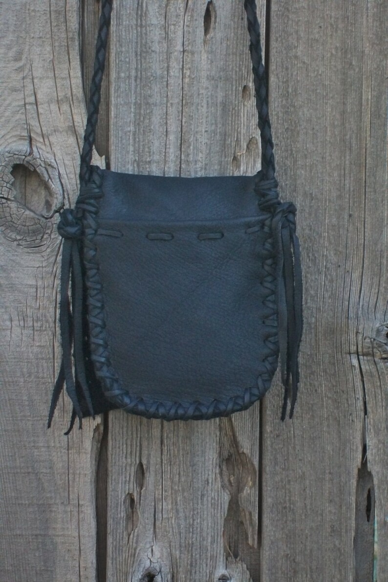 K&ouml;nnte beinhalten: Eine schwarze Ledertasche mit geflochtenem Trageriemen und Fransen. Die Tasche hat einen Klappverschluss und h&auml;ngt an einer Holzoberfl&auml;che.