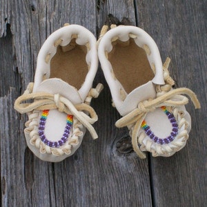May include: A pair of white leather moccasins with purple and rainbow beaded trim. The moccasins are on a wooden surface.