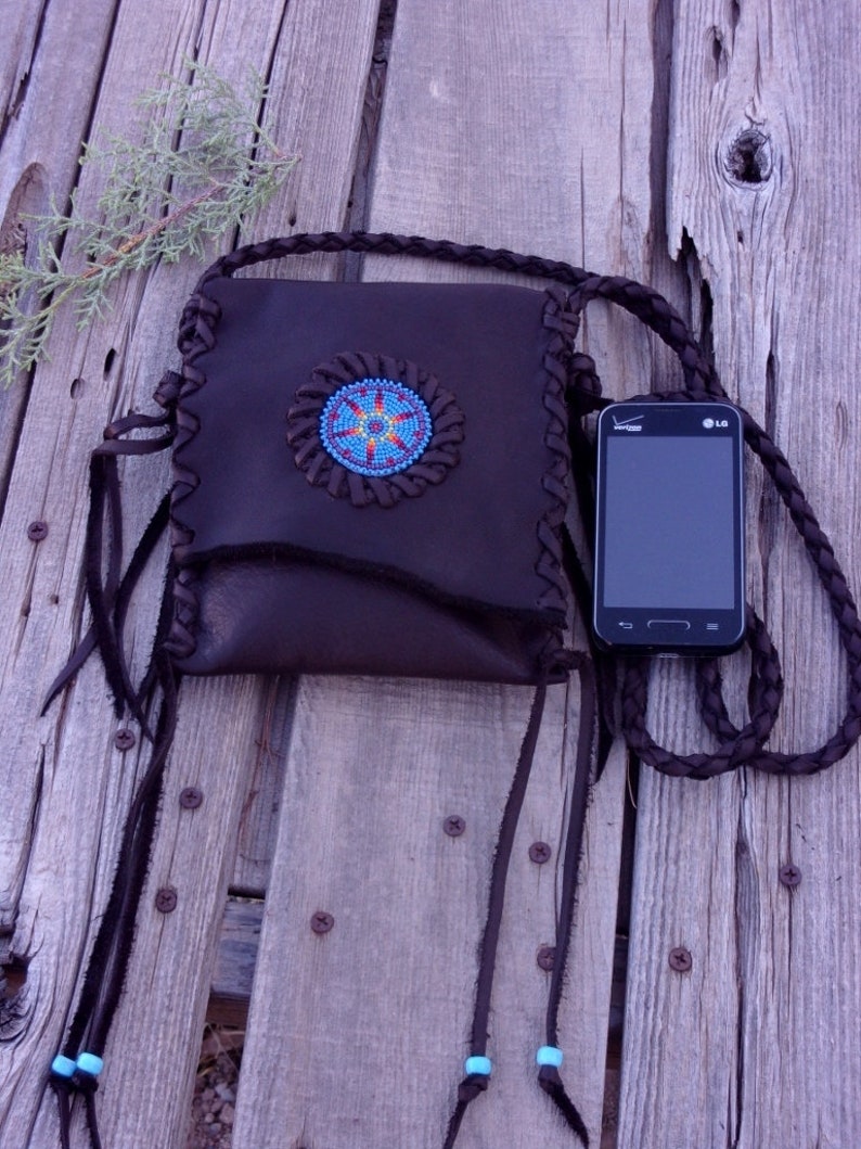 May include: A brown leather purse with a beaded design and fringe. The purse has a long strap and is decorated with turquoise beads. A black cell phone with the LG logo is visible in the image.