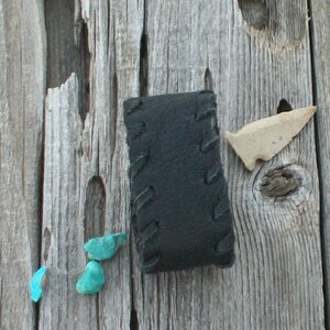 May include: A black leather pouch with stitching detail, three turquoise stones, and a small arrowhead on a weathered wooden surface.
