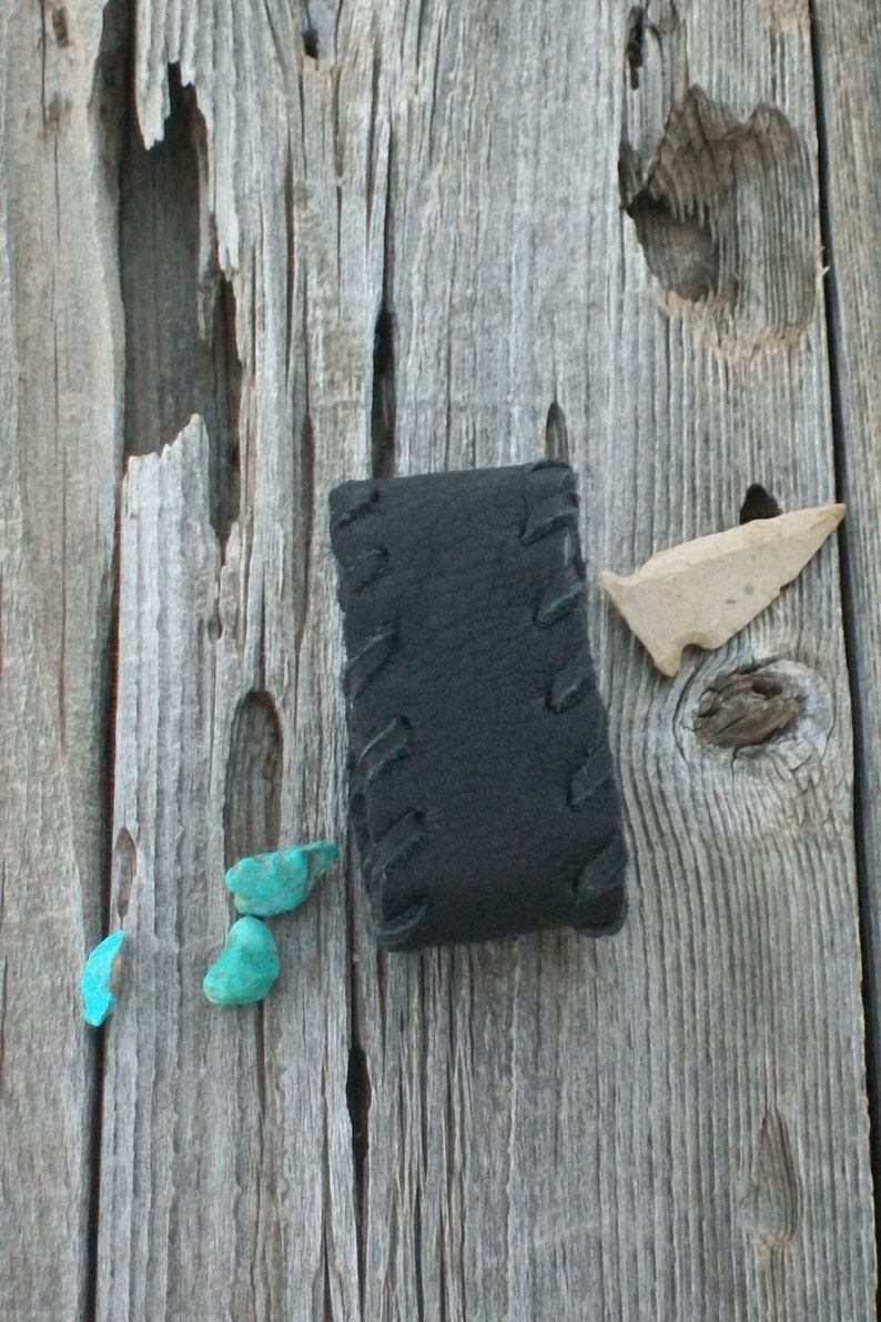 May include: A black leather pouch with stitching detail, three turquoise stones, and a small arrowhead on a weathered wooden surface.