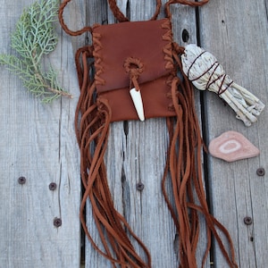 Pu&ograve; includere: Una borsa in pelle marrone con frange e un ciondolo in osso. La borsa &egrave; appesa a una tracolla in pelle intrecciata. C'&egrave; un mazzo di salvia bianca e una piccola pietra accanto alla borsa.