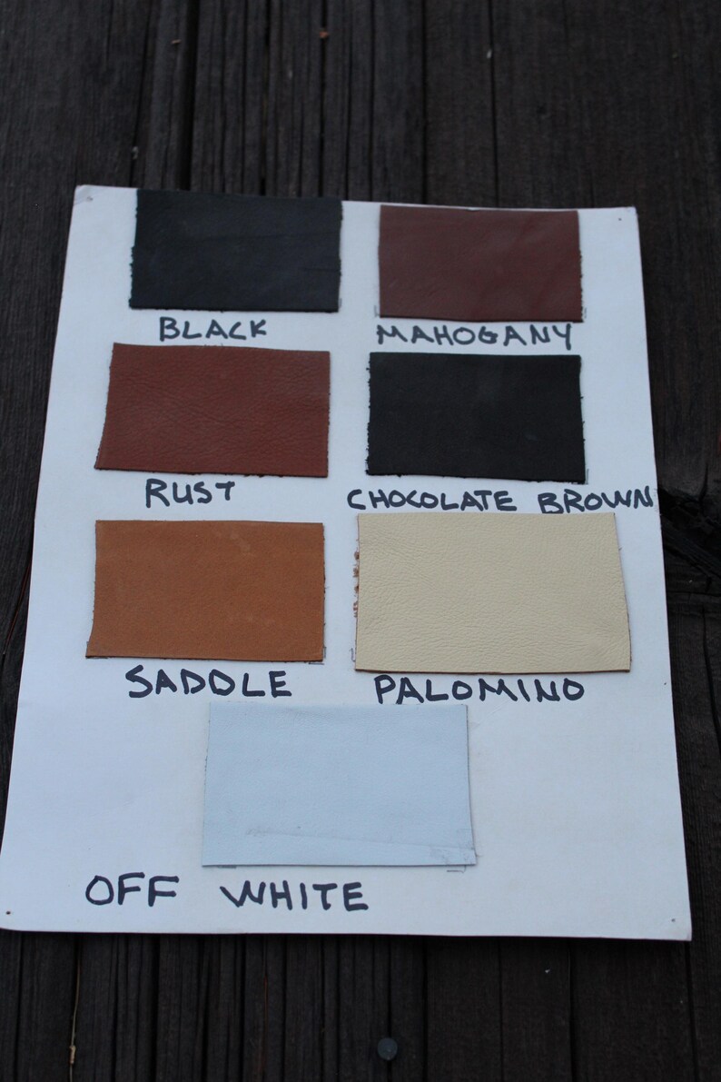 May include: A white display board featuring seven rectangular leather swatches in various colors labeled Black, Mahogany, Rust, Chocolate Brown, Sadole, Palomino, and Off White, with each color name written in black ink below its respective swatch.