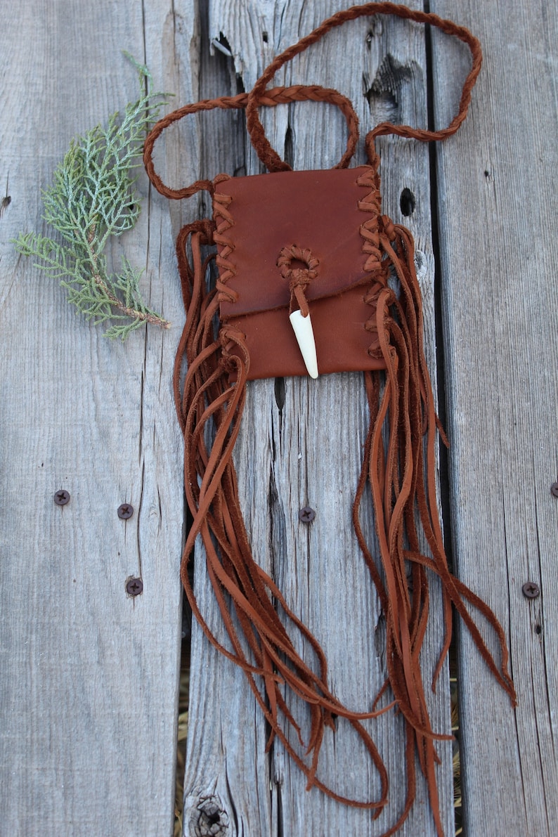 Pu&ograve; includere: Una piccola borsa in pelle marrone con una frangia e un accento in osso. La borsa &egrave; appesa a una cinghia in pelle intrecciata.