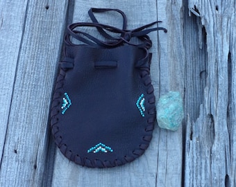 May include: A small brown leather pouch with a drawstring closure. The pouch is decorated with turquoise and white beadwork in a geometric pattern. The pouch is sitting on a wooden surface.