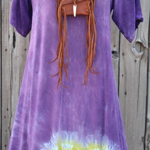 Pu&ograve; includere: Un vestito viola tie-dye con una borsa di pelle marrone con frange appesa a un cordino di pelle intorno al collo.