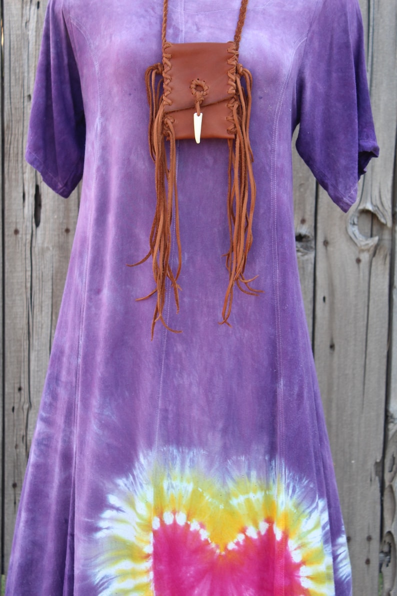 Pu&ograve; includere: Un vestito viola tie-dye con una borsa di pelle marrone con frange appesa a un cordino di pelle intorno al collo.