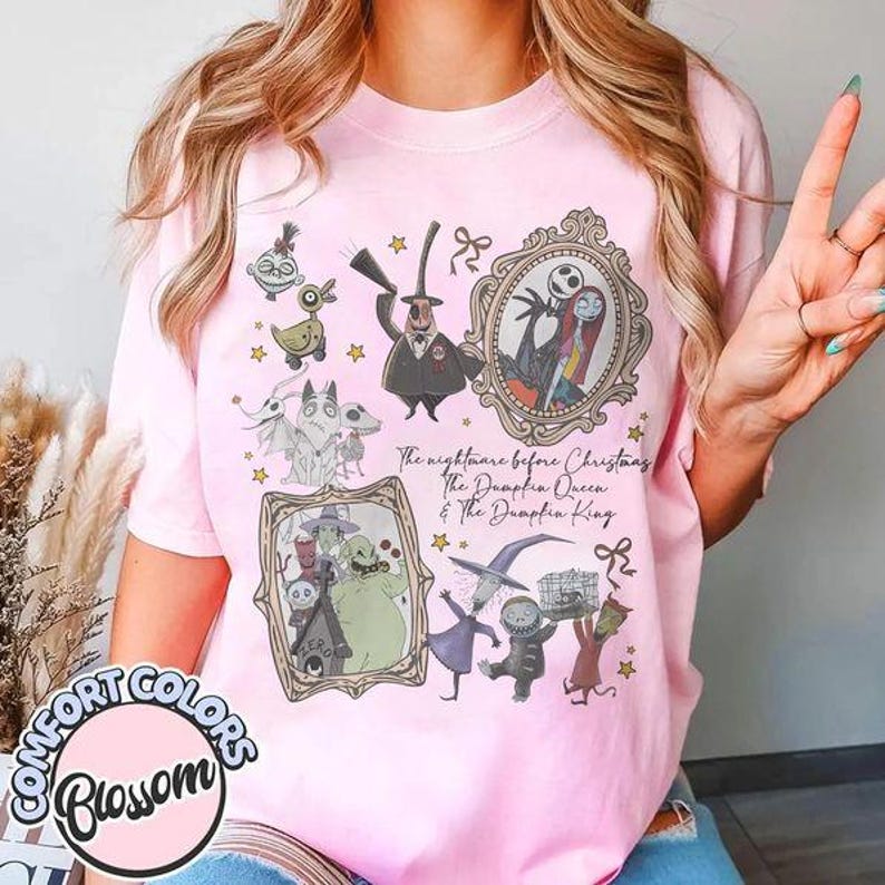 May include: A light pink t-shirt featuring a graphic print of characters from The Nightmare Before Christmas. The design includes Jack Skellington, Sally, Oogie Boogie, and Zero, with the text The Nightmare Before Christmas The Pumpkin Queen & The Pumpkin King.