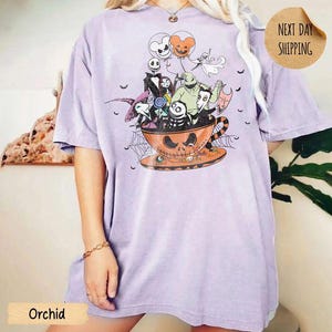 Pode incluir: Uma t-shirt roxo-claro com uma estampa gr&aacute;fica de personagens de O Estranho Mundo de Jack dentro de uma grande ch&aacute;vena de ch&aacute; laranja em forma de ab&oacute;bora. O design inclui bal&otilde;es, teias de aranha e v&aacute;rias figuras assustadoras agrupadas.