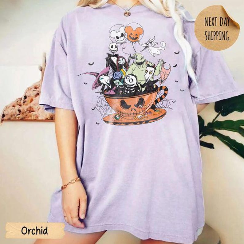 Pode incluir: Uma t-shirt roxo-claro com uma estampa gr&aacute;fica de personagens de O Estranho Mundo de Jack dentro de uma grande ch&aacute;vena de ch&aacute; laranja em forma de ab&oacute;bora. O design inclui bal&otilde;es, teias de aranha e v&aacute;rias figuras assustadoras agrupadas.