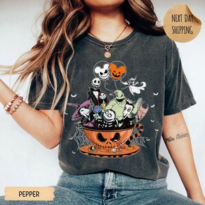 Pode incluir: Uma t-shirt cinzento-carv&atilde;o com uma impress&atilde;o gr&aacute;fica de v&aacute;rias personagens animadas de O Estranho Mundo de Jack dentro de uma grande ch&aacute;vena de ch&aacute; laranja em forma de ab&oacute;bora, acompanhada por bal&otilde;es flutuantes e teias de aranha.
