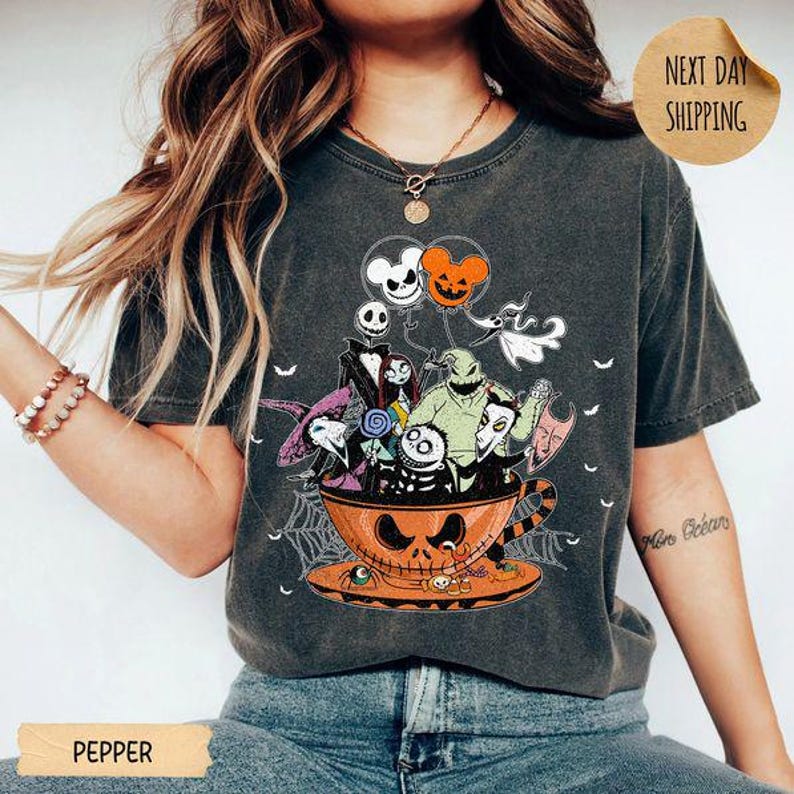 Pode incluir: Uma t-shirt cinzento-carv&atilde;o com uma impress&atilde;o gr&aacute;fica de v&aacute;rias personagens animadas de O Estranho Mundo de Jack dentro de uma grande ch&aacute;vena de ch&aacute; laranja em forma de ab&oacute;bora, acompanhada por bal&otilde;es flutuantes e teias de aranha.