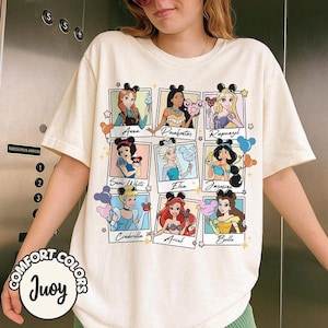 May include: Cream-colored t-shirt featuring nine Disney princesses in polaroid-style frames. Each princess is depicted with a Mickey Mouse ear headband and holding a themed item. The shirt has the words "COMFORT COLORS" and "Juoy" printed on it.