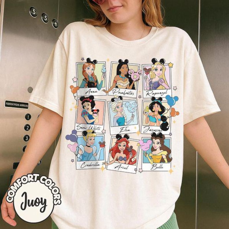 May include: Cream-colored t-shirt featuring nine Disney princesses in polaroid-style frames. Each princess is depicted with a Mickey Mouse ear headband and holding a themed item. The shirt has the words "COMFORT COLORS" and "Juoy" printed on it.