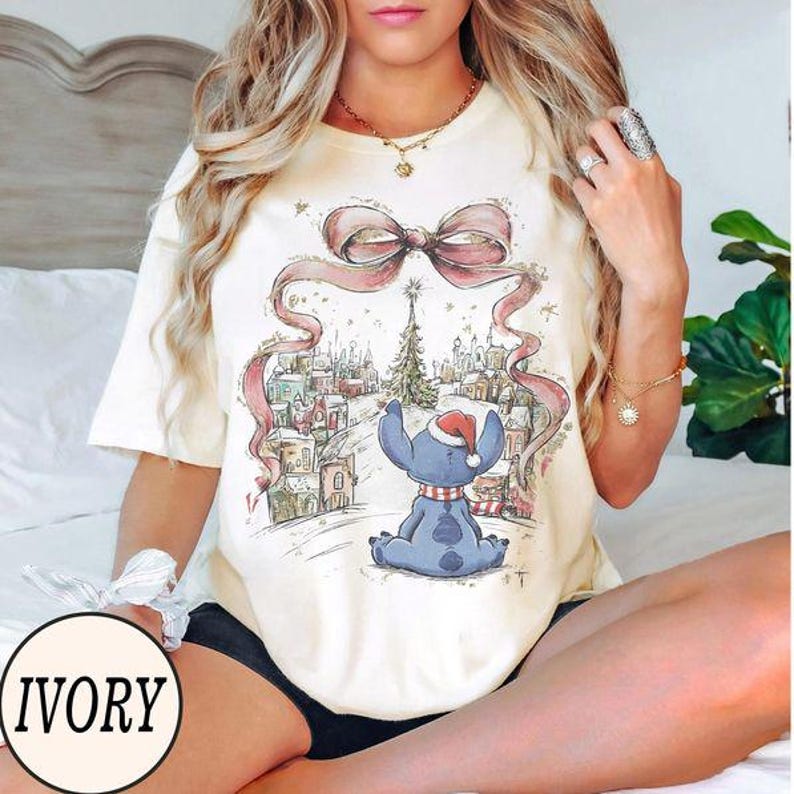 Puede incluir: Una camiseta de manga corta color marfil con un estampado gr&aacute;fico de una criatura azul con un gorro de Pap&aacute; Noel rojo, sentada frente a una escena de pueblo nevado bajo un gran lazo rosa y un &aacute;rbol de Navidad.