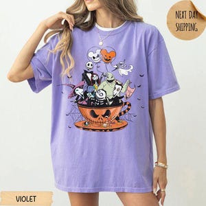 Pode incluir: Uma t-shirt violeta de manga curta com uma impress&atilde;o gr&aacute;fica de personagens de O Estranho Mundo de Jack sentados dentro de uma grande ch&aacute;vena de ch&aacute; laranja. O design inclui Jack Skellington, Sally, Oogie Boogie e Zero com bal&otilde;es.