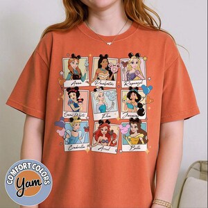 May include: Orange Comfort Colors t-shirt featuring nine Disney princesses in framed portraits. Each princess holds a Mickey Mouse-shaped ice cream. The shirt has the Comfort Colors and Yam logos on the bottom left.
