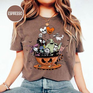 May include: Espresso-colored t-shirt featuring a Halloween-themed graphic. The design includes characters from The Nightmare Before Christmas inside a jack-o'-lantern teacup, with Mickey Mouse-shaped balloons. The shirt has a crew neck and short sleeves.