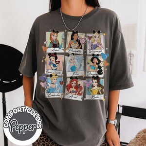 May include: A gray Comfort Colors t-shirt featuring nine Disney princesses in framed portraits. Each princess is wearing a mouse ear headband. The princesses' names are printed below their portraits. The shirt has the Comfort Colors Pepper logo.