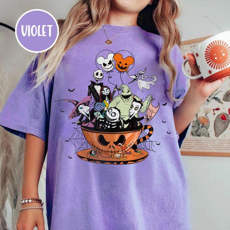 May include: A violet t-shirt featuring a Halloween-themed graphic. The design includes characters from "The Nightmare Before Christmas" inside an orange teacup with a skull face. The shirt also has the word "VIOLET" in a purple circle.
