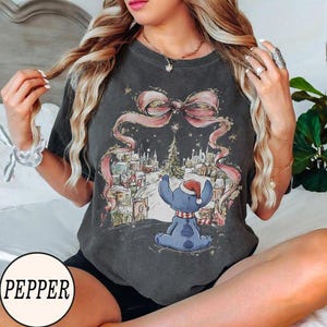 Puede incluir: Una camiseta de manga corta gris carb&oacute;n con un estampado gr&aacute;fico de una criatura azul con gorro de Pap&aacute; Noel, sentada frente a un pueblo nevado bajo un gran lazo rosa. La camiseta tiene cuello redondo y un acabado de estilo vintage.
