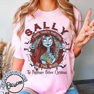 以下が含まれることがあります： 「The Nightmare Before Christmas」のサリーが描かれたライトピンクのTシャツ。デザインには「I SENSE THERE'S SOMETHING IN THE WIND」、「EST. 1993」、「The Nightmare Before Christmas」という文字と、クモの巣やコウモリのイラストが含まれています。