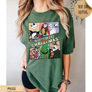 May include: A moss green short-sleeve t-shirt featuring a graphic print of characters from The Nightmare Before Christmas. The design includes panels of Jack Skellington, Sally, Zero, Oogie Boogie, and other figures with the text Nightmare Before Christmas.