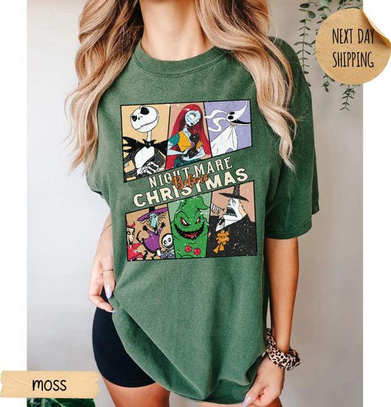 May include: A moss green short-sleeve t-shirt featuring a graphic print of characters from The Nightmare Before Christmas. The design includes panels of Jack Skellington, Sally, Zero, Oogie Boogie, and other figures with the text Nightmare Before Christmas.