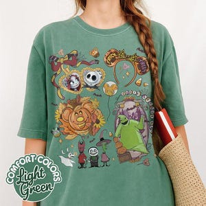 Pode incluir: Uma t-shirt verde-s&aacute;lvia com uma impress&atilde;o gr&aacute;fica de personagens e &iacute;cones de O Estranho Mundo de Jack, incluindo Jack Skellington, Sally e Oogie Boogie, juntamente com elementos de outono como ab&oacute;boras e doces de milho.
