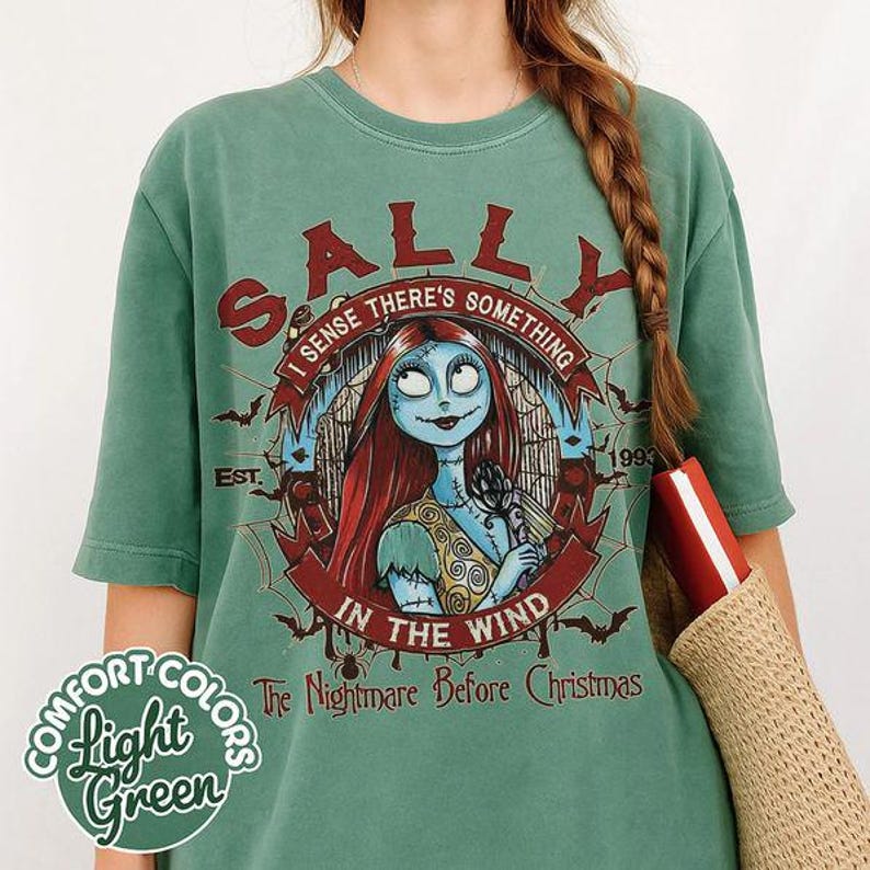 以下が含まれることがあります： セージグリーンのTシャツで、『ナイトメアー・ビフォア・クリスマス』のキャラクター、サリーのグラフィックが描かれています。デザインには「SALLY」、「I SENSE THERE'S SOMETHING IN THE WIND」、「EST. 1993」、「The Nightmare Before Christmas」という文字とコウモリが含まれています。