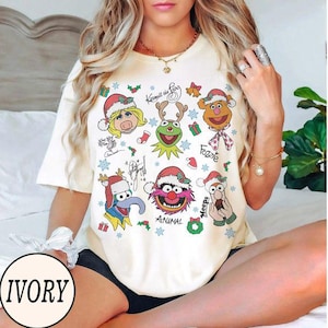 May include: Ivory-colored t-shirt featuring a festive design with various Muppet characters wearing Santa hats. The shirt includes the text "Kermit the Frog", "Fozzie", and "Animal". Snowflakes and gift box graphics are also present.