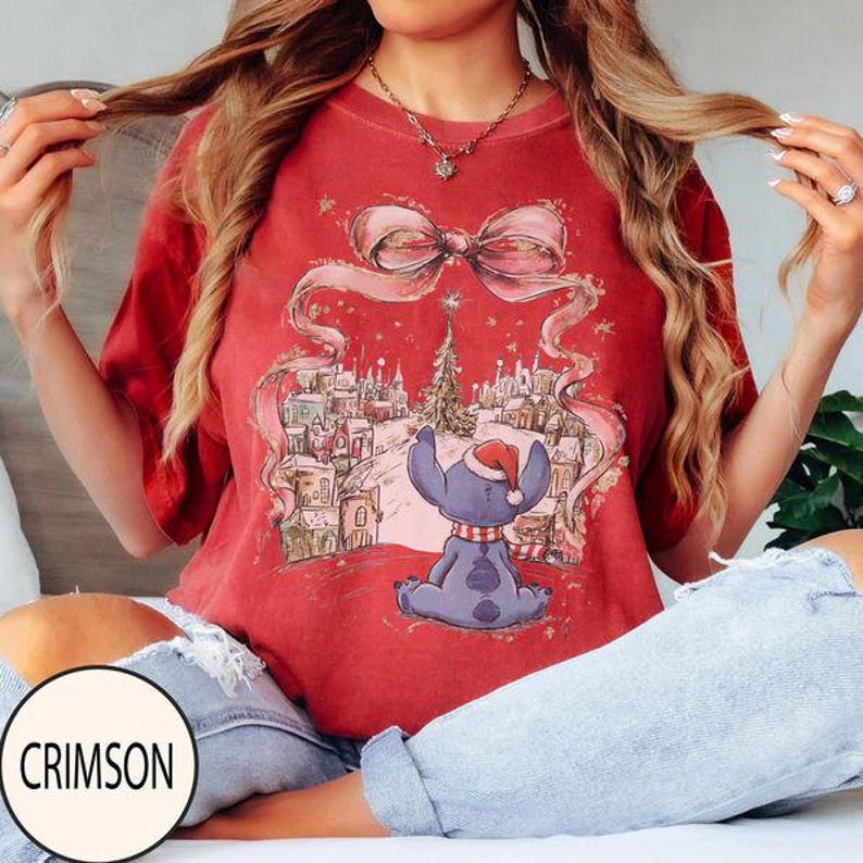 Puede incluir: Una camiseta de manga corta color rojo carmes&iacute; con un estampado gr&aacute;fico de una criatura azul con gorro de Pap&aacute; Noel, sentada frente a un pueblo nevado con un &aacute;rbol de Navidad, enmarcado por un gran dise&ntilde;o de lazo rosa en el frente.