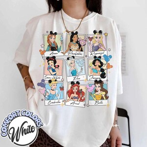 May include: White Comfort Colors t-shirt featuring nine Disney princesses in framed portraits. Each princess is depicted with a cotton candy treat and wearing mouse ears. The princesses include Anna, Pocahontas, Rapunzel, Snow White, Elsa, Jasmine, Cinderella, Ariel, and Belle.