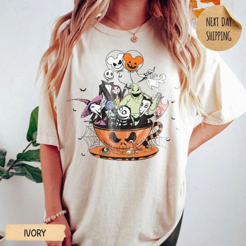 Pode incluir: Uma t-shirt cor de marfim com uma impress&atilde;o gr&aacute;fica de v&aacute;rias personagens de O Estranho Mundo de Jack dentro de uma grande ch&aacute;vena de ch&aacute; laranja. O design inclui Jack Skellington, Sally, Oogie Boogie e Zero, com bal&otilde;es e teias de aranha.