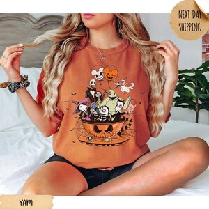 Pode incluir: Uma t-shirt de gola redonda laranja com uma impress&atilde;o gr&aacute;fica de personagens de O Estranho Mundo de Jack dentro de uma grande ch&aacute;vena de ch&aacute; em forma de ab&oacute;bora. O design inclui Jack Skellington, Sally, Zero e Oogie Boogie com bal&otilde;es.
