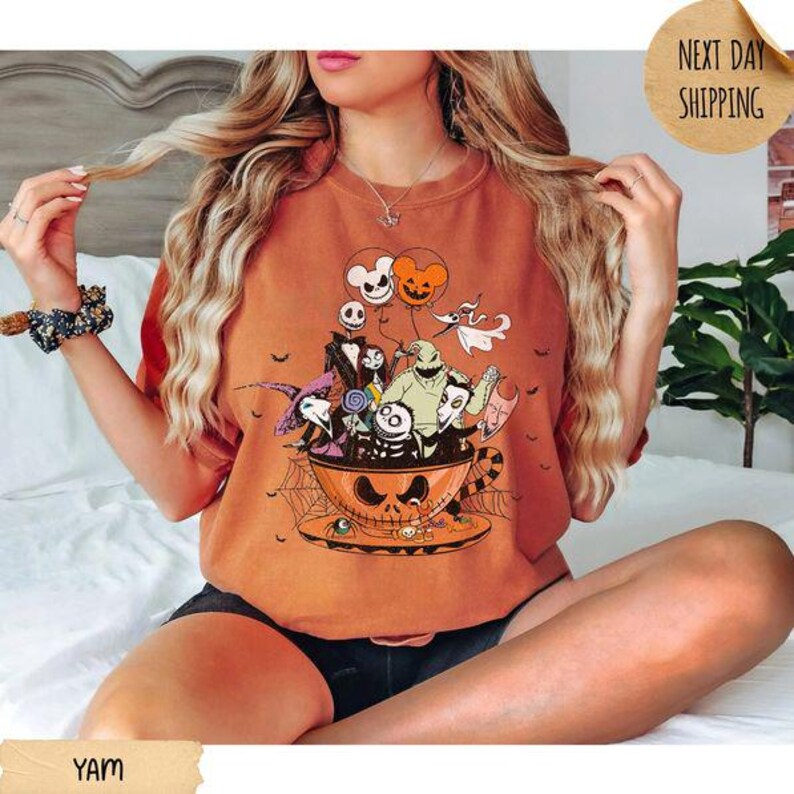 Pode incluir: Uma t-shirt de gola redonda laranja com uma impress&atilde;o gr&aacute;fica de personagens de O Estranho Mundo de Jack dentro de uma grande ch&aacute;vena de ch&aacute; em forma de ab&oacute;bora. O design inclui Jack Skellington, Sally, Zero e Oogie Boogie com bal&otilde;es.