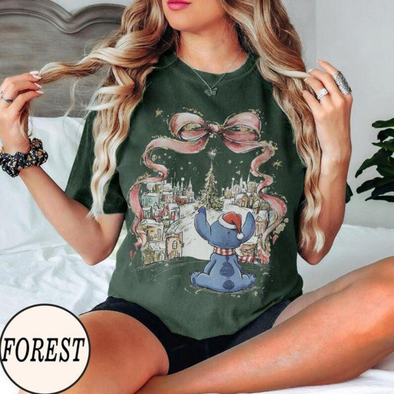 Puede incluir: Una camiseta de manga corta color verde bosque con un estampado gr&aacute;fico de un personaje azul con gorro de Pap&aacute; Noel, sentado frente a un pueblo nevado. Un gran lazo rosa est&aacute; colocado sobre la parte superior de la ilustraci&oacute;n.
