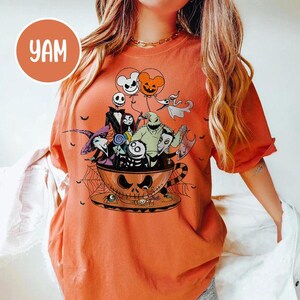 May include: Orange t-shirt featuring a Halloween-themed graphic. The design includes characters from "The Nightmare Before Christmas" inside a teacup with a jack-o'-lantern face. The shirt also has the text "YAM" in a circle.