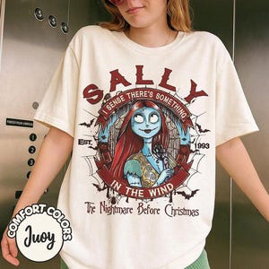 以下が含まれることがあります： クリーム色の半袖Tシャツで、『ナイトメアー・ビフォア・クリスマス』のサリーのグラフィックが描かれています。デザインには「SALLY」、「I SENSE THERE'S SOMETHING IN THE WIND」、「EST. 1993」、「The Nightmare Before Christmas」という文字とコウモリのイラストが含まれています。