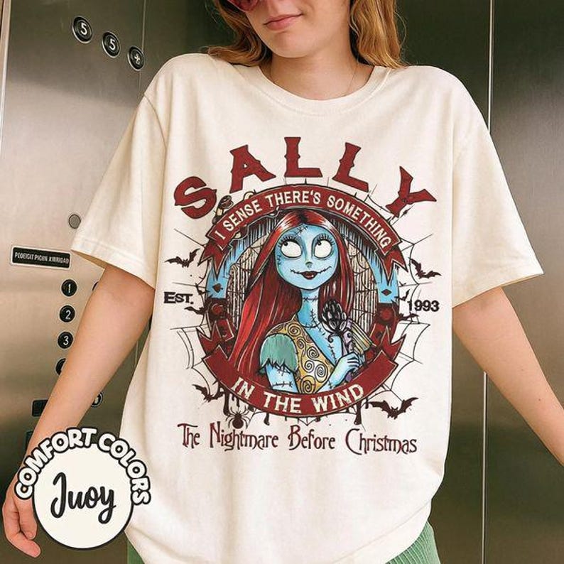 以下が含まれることがあります： クリーム色の半袖Tシャツで、『ナイトメアー・ビフォア・クリスマス』のサリーのグラフィックが描かれています。デザインには「SALLY」、「I SENSE THERE'S SOMETHING IN THE WIND」、「EST. 1993」、「The Nightmare Before Christmas」という文字とコウモリのイラストが含まれています。