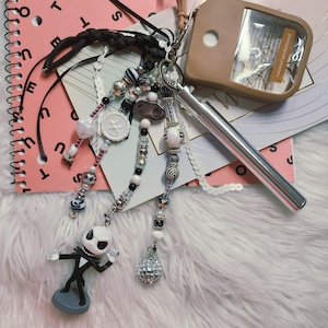 Puede incluir: Un accesorio de llavero que presenta una figura del personaje Jack Skellington, un bol&iacute;grafo de metal plateado y un soporte para desinfectante de manos color canela etiquetado como coconut sandalwood. Incluye varias hileras de cuentas con patrones.