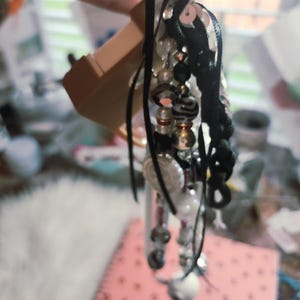 Puede incluir: Un adorno colgante decorativo que presenta una disposici&oacute;n vertical de cuentas negras, blancas y plateadas met&aacute;licas, intercaladas con cintas negras, lentejuelas y elementos esf&eacute;ricos estampados, suspendido de un gancho de exhibici&oacute;n de pl&aacute;stico beige.