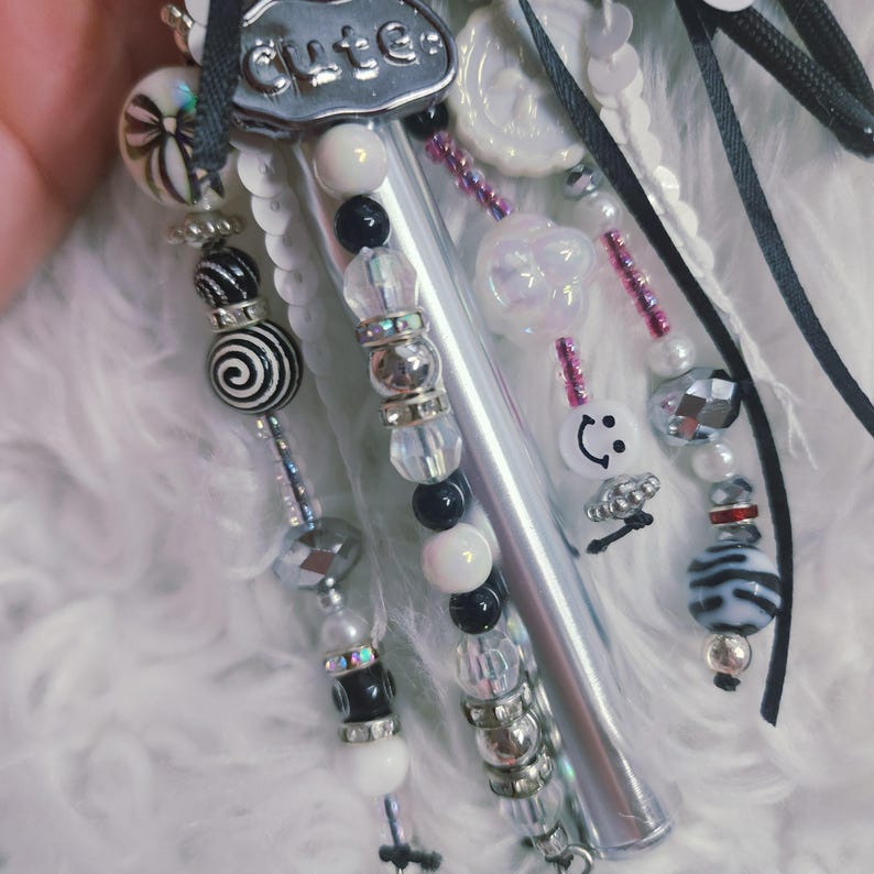Puede incluir: Un bol&iacute;grafo decorativo con cuerpo plateado rematado por un dije de metal grabado con la palabra cute. El bol&iacute;grafo est&aacute; adornado con m&uacute;ltiples hilos de cuentas en tonos negros, blancos e iridiscentes, incluyendo cuentas en espiral, con caras sonrientes y flores, acentuado con cintas negras y lentejuelas blancas.