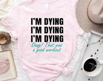 May include: A light pink t-shirt with the text "I'M DYING" repeated three times in black, followed by "Dang! That was a good workout" in teal. The shirt is styled with a hat, tray with coffee, and a book.