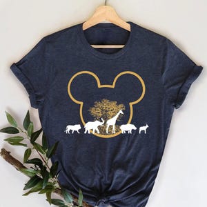 May include: Navy blue t-shirt featuring a gold outline of Mickey Mouse ears with a tree and safari animals inside. The design includes a giraffe, elephant, and other animals in white. The shirt is hanging on a wooden hanger.