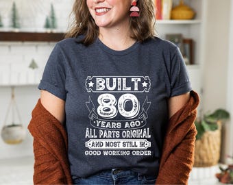 May include: A dark gray t-shirt with the text "BUILT 80 YEARS AGO ALL PARTS ORIGINAL AND MOST STILL IN GOOD WORKING ORDER" in white. The shirt is a crew neck and has rolled-up sleeves.