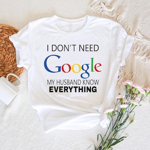 May include: White t-shirt with the text "I DON'T NEED Google MY HUSBAND KNOW EVERYTHING" in black, blue, red, yellow, and green. The shirt is knotted at the bottom. The background includes a woven basket, a white mug, and flowers.