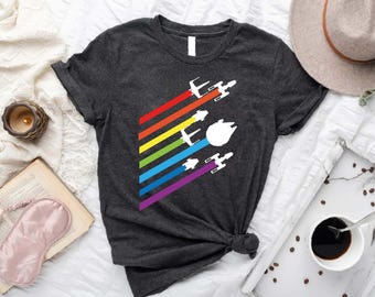 May include: Dark gray t-shirt featuring a colorful graphic of spaceships and a spaceship with a white background. The design includes a rainbow of colors and a white spaceship. The shirt is knotted at the waist.